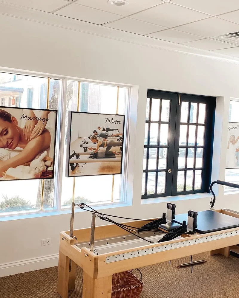 A Pilates reformer beside tall windows, with framed massage and Pilates prints on the wall