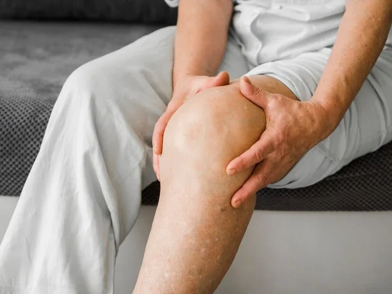 An older person seated on a bed with both hands wrapped around one knee