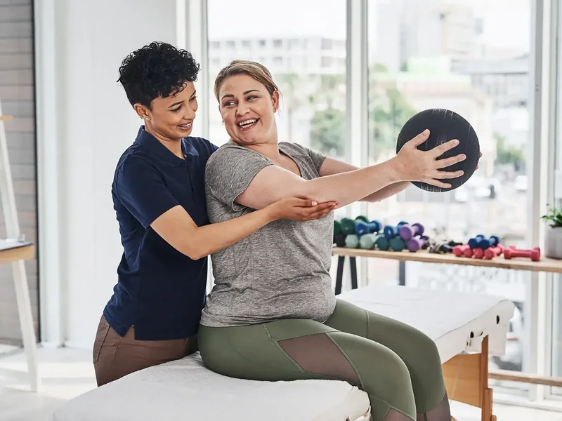 A therapist guiding a seated woman's arms as she holds a weighted ball out in front of her